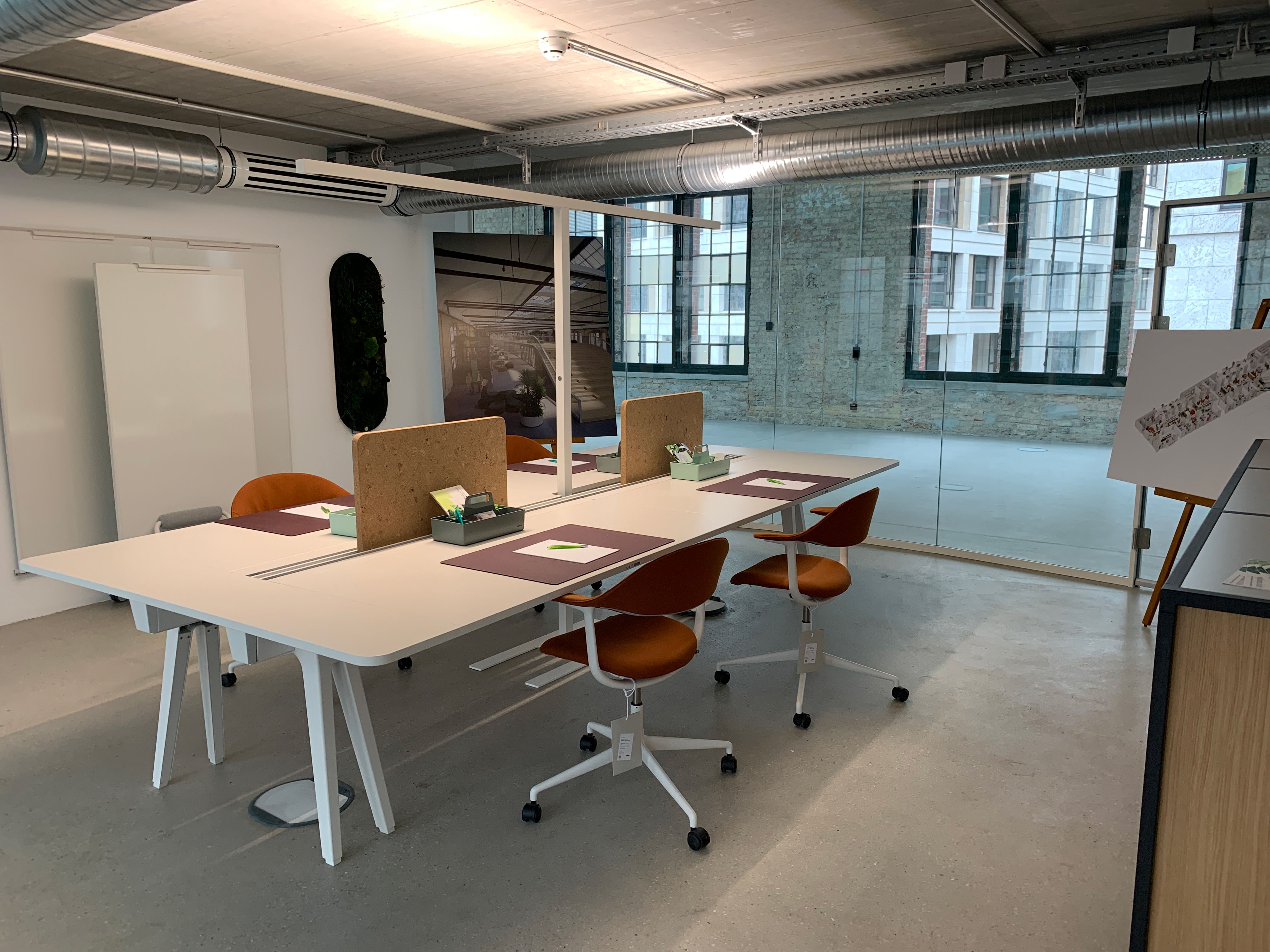 Bright workplace with white desks, orange chairs, and a view through a glass wall in an industrial setting.