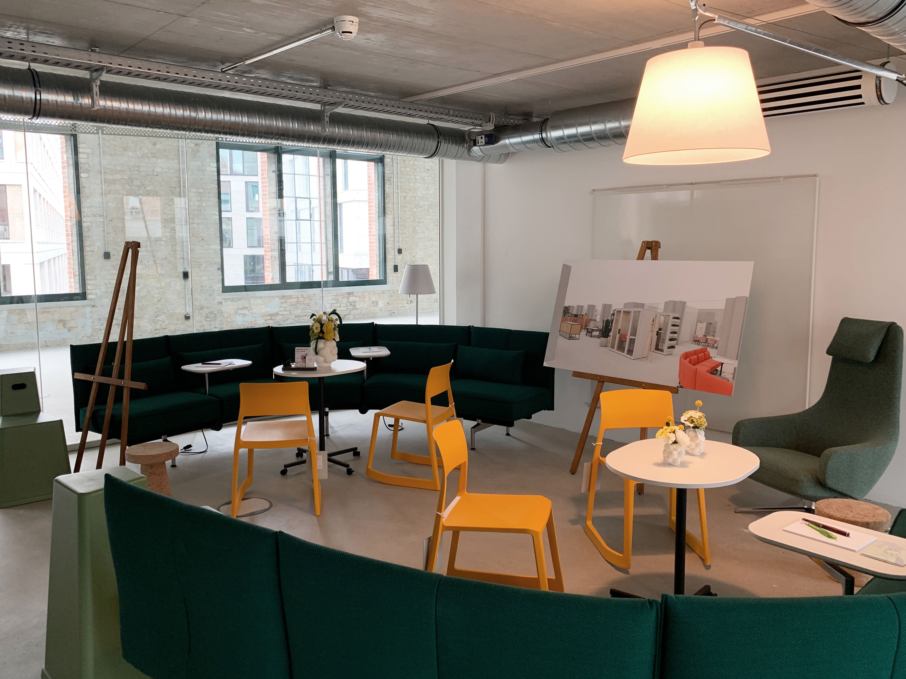 Comfortable lounge with green sofas and yellow chairs, furnished meeting area with model boards and floor lamps.