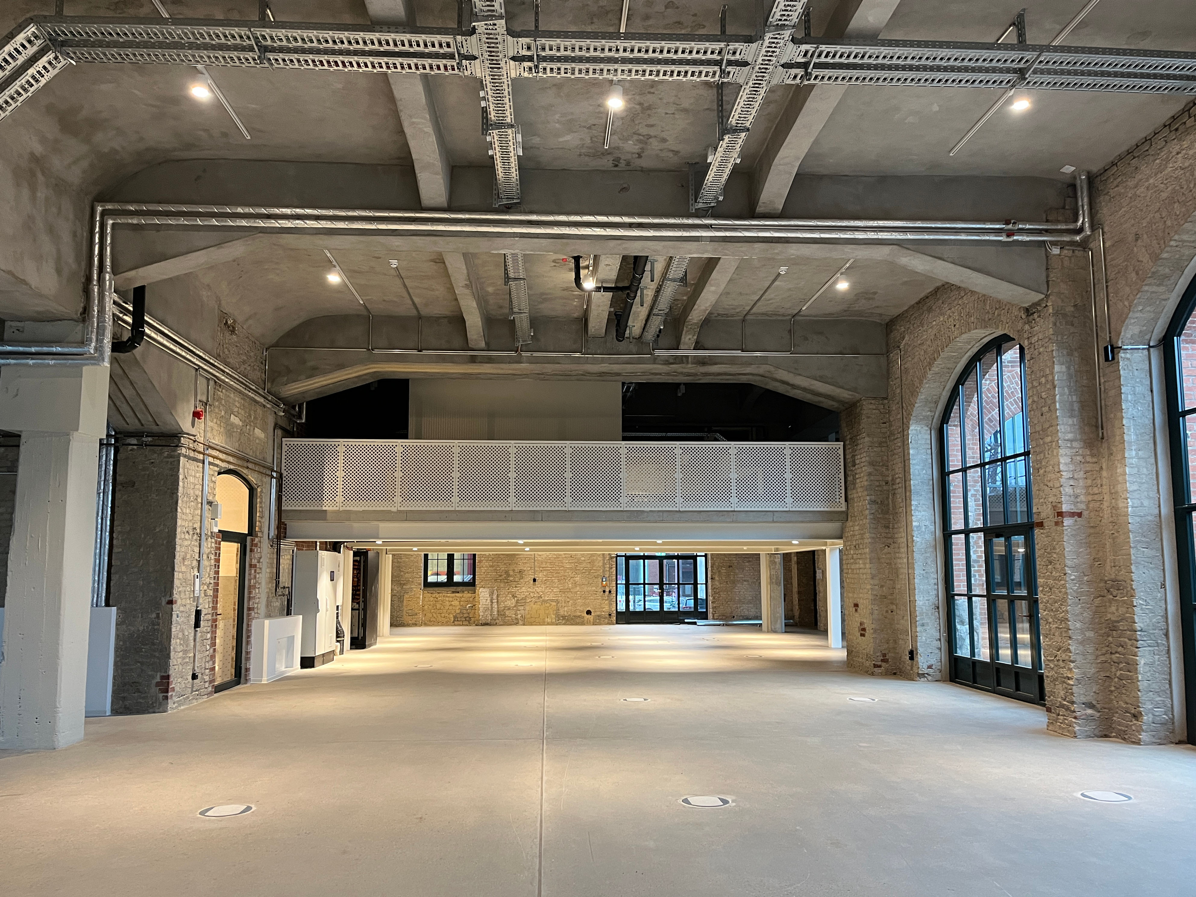 Der Baufortschritt im Postbahnhof Berlin zeigt die Abgangspackkammer im Erdgeschoss als weiten, leeren Raum mit Betonboden, gemauerten Pfeilern, Rundbögen und sichtbaren technischen Installationen an der Decke.