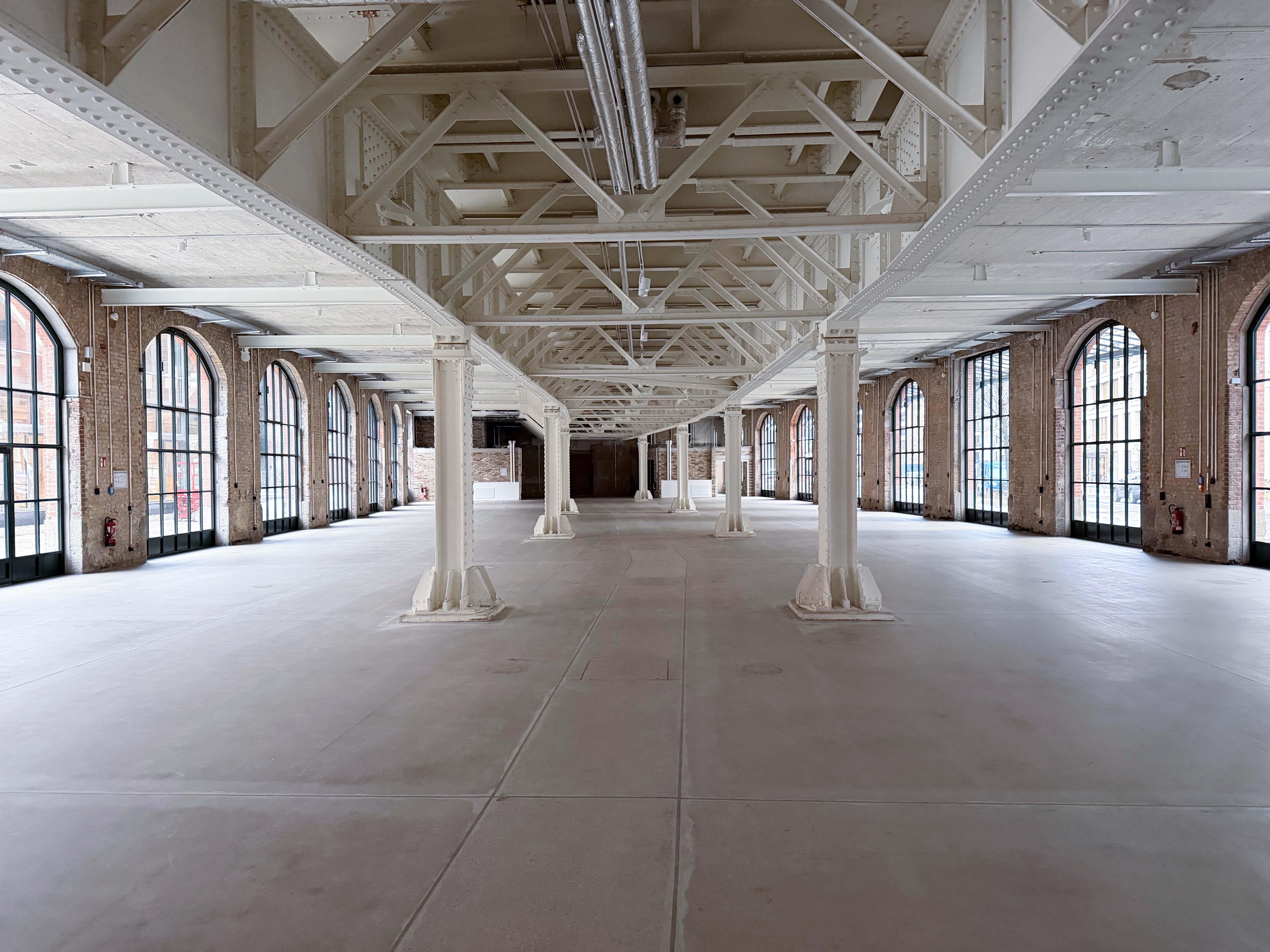 Der Baufortschritt im Postbahnhof Berlin zeigt die Eingangspackkammer im Erdgeschoss mit freistehenden Stützen, hohen Decken und umlaufenden Fensterfronten.