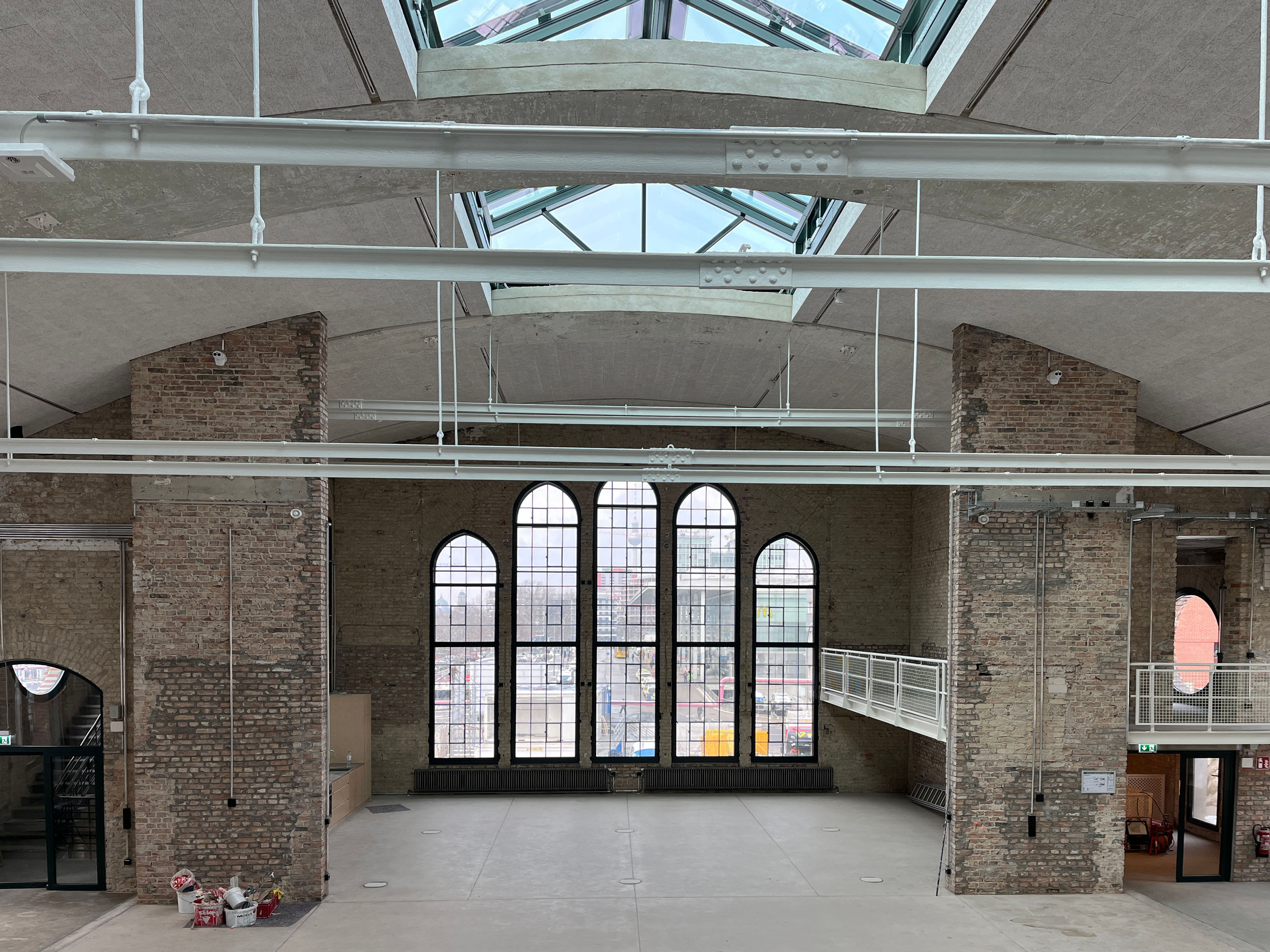 The image shows the construction progress at Postbahnhof Berlin inside the entrance packing hall with historic arched windows and exposed roof structure.